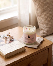 A glass jar candle with a pink label reading 'Blossom's Bouquet' and 'Rosemary Scott' sits on a small wooden table beside a book and dried flowers in a softly lit room.