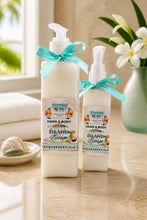 Two bottles of hand and body lotion with blue ribbons on a wooden surface, with a vase of flowers and towels in the background.
