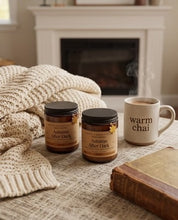 Two amber glass jars labeled 'Autumn After Dark' are styled on a table with a cream knit throw blanket, a white mug reading 'warm chai', and an old book in front of a fireplace.