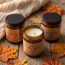 Three amber glass candles with black lids and tan labels reading 'First Sip Of Fall' are arranged on a rustic wooden surface with scattered autumn leaves and a cream knit blanket in the background.