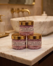 Three jars of whipped soap scrub stacked on a marble bathroom counter, with a marble sink and gold faucet blurred in the background. The labels read 'Whipped Soap Scrub' and 'A Rose Remembered'.