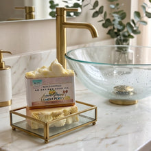 A bar of artisan soap labeled 'Lemon Poppy' by Benjamins Handmade sits on a gold-framed glass tray on a white marble bathroom counter next to a gold faucet and a clear glass bowl.