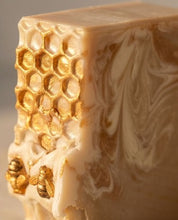 Close-up of a decorative bar of soap with a honeycomb pattern, gold leaf details, and gold bee embellishments on a light beige background.