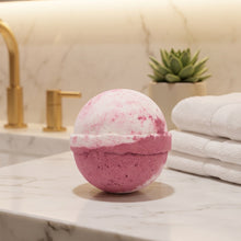 A pink and white marbled bath bomb sits on a white marble bathroom countertop next to folded white towels, with a gold faucet and a small potted succulent in the background.