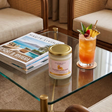 A glass jar candle with a gold lid and pink label sits on a glass-top table beside a tall cocktail garnished with fruit, with two woven armchairs and a magazine in the background.