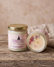 Two Bridgerton's Bouquet scented soy candles by RCOC Essentials on a rustic wooden surface. One candle is upright with a gold lid and a pink label, and the other is lying down showing the cream wax with colorful confetti and glitter.