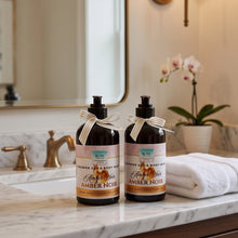 Two amber glass shower gel and body wash bottles with cream-colored labels and ribbon bows, set on a marble bathroom counter next to a folded white towel.