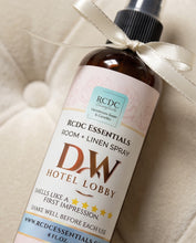 A bottle of RCDC Essentials Room + Linen Spray by DW Hotel Lobby, wrapped in a cream satin ribbon, set against a soft, textured cream fabric background.