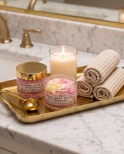 A bathroom scene showing a gold tray holding a jar of whipped soap scrub, a lit candle, rolled towels, and a small gold cup on a marble countertop near a sink.
