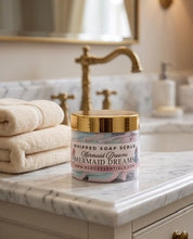 A jar of whipped soap scrub labeled 'Mermaid Dreams' sits on a marble bathroom counter beside folded cream towels, with a gold faucet and softly blurred bathroom background.