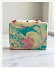A rectangular bar of marbled soap with swirls of teal, pink, and cream, topped with small pink flower decorations, standing on a light marble surface.