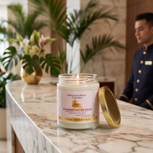 A lit candle in a clear glass jar sits on a marble counter with its gold lid beside it. A hotel-style lobby with palm leaves and a uniformed staff member is softly blurred in the background.