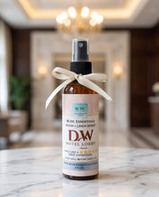 A bottle of RCDC Essentials Room + Linen Spray by DW Hotel Lobby, featuring an amber glass bottle, black spray cap, and a cream ribbon bow tied around the neck, set on a marble countertop with a blurred hotel lobby background.