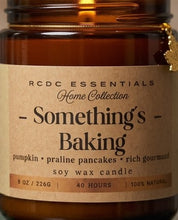 An amber glass jar candle with a kraft paper label reading 'RCDC ESSENTIALS Home Collection - Something's Baking' and 'soy wax candle' on a warm brown background.