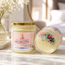 Two glass jar candles on a light marble surface in a softly lit room. One candle is upright with a pink label reading 'She's Beautiful' and 'RCDC Essentials,' and the other is tipped on its side showing pale yellow wax topped with multicolored glitter.