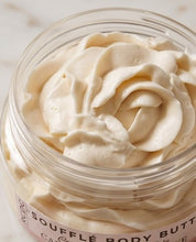 A close-up of a clear jar filled with whipped, creamy off-white body butter, with a partial label reading 'SOUFFLÉ BODY BUTTER' on a light marble-like background.