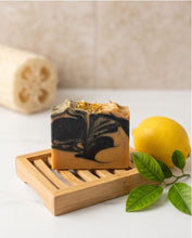 A rectangular bar of marbled soap with black and warm tan swirls sits on a small bamboo soap dish. A yellow lemon and green leaves are placed beside it, with a loofah blurred in the background against a light beige tiled wall.