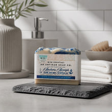 A bar of artisan soap from RCDC Essentials, labeled 'Anchors Aweigh', displayed on a dark slate tray in a modern bathroom setting with a soap dispenser and tiled wall in the background.