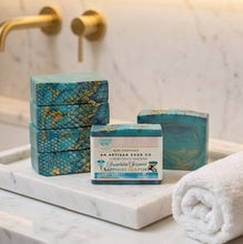 Three teal and blue patterned soap bars with labels from An Artisan Soap Co. arranged on a white marble tray next to a rolled white towel, set against a marble bathroom wall with gold faucets.