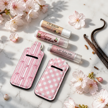 Three lip balm tubes and two pink fabric lip balm cases arranged on a white marble surface with cherry blossoms and vanilla beans. The balm tubes include Vanilla, Cherry Bomb, and Mint flavors, and the cases feature pink-and-white gingham and pink-and-white vertical stripes.