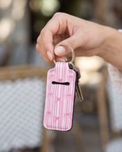 A hand holding a pink and white striped keychain wallet with a black coin slot and a gold key ring, set against a blurred outdoor patio background.