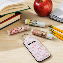 A close-up tabletop scene showing several lip balm tubes and a patterned key fob case on a light wood desk, with books, an apple, pencils, and a notebook around them. The lip balm tubes appear to be the products being sold: one Cherry Bomb, one Vanilla Vanilla, and one RDG lip balm.