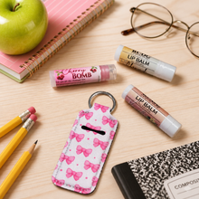 A pink and white bow-patterned keychain money box with a metal key ring, styled on a light wood desk with school supplies including a green apple, notebooks, pencils, and lip balm tubes.
