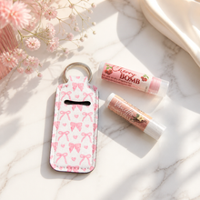 A pink and white patterned keychain money pouch with a coin slot, styled on a white marble surface next to two lip balm tubes labeled Cherry Bomb and Willy Rose, with soft pink flowers in the background.