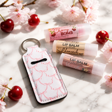Two lip balm tubes and a white keychain pouch with pink bow patterns are arranged on a white marble surface with cherry blossoms and cherries around them. The lip balm labels show the brands Nicole Lee and Vanessa Goyder.