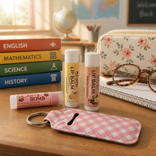 A styled desk scene with several lip balm tubes and a pink-and-white gingham key fob on a wooden table, alongside books, glasses, a notebook, and a floral pouch in the background.