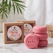 Pink bath bombs and their kraft paper packaging boxes on a white marble surface with a green plant leaf in the background. The brand name RCDC is visible on the labels and embossed on the solid shampoo bar.