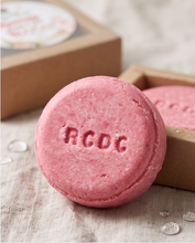 A round pink solid shampoo bar embossed with the brand name 'RCDC' in the center, resting on a textured beige fabric surface next to its brown kraft paper packaging box.