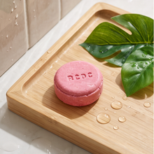 A pink round solid shampoo bar embossed with the brand name 'noco' sits on a light bamboo tray, styled with green monstera leaves and water droplets on a marble surface.