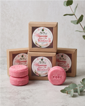 Three small kraft paper boxes with round floral labels are arranged behind two pink round solid shampoo bars, with one pink bar placed in front. The product branding reads RCDC and the label text includes Strawberry and Blooms.