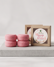 Four round pink solid shampoo bars are arranged in two stacks beside two kraft gift boxes, one with a round label that reads 'Rose & Bloom' and 'Scrub & Soak.' The setup is photographed on a light gray stone slab against a soft light gray background.