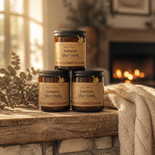 Three amber glass candles labeled 'Autumn After Dark' by Beige Essentials are stacked on a rustic wooden mantel in front of a cozy fireplace, with a cream knit throw and eucalyptus branches nearby.