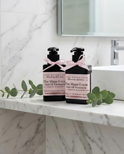Two black hair oil treatment bottles with pink ribbon bows and white labels reading 'The Mane Event' and 'Traveling Hair Oil' by Kloe Blisterman, styled on a white marble countertop with eucalyptus leaves in a bathroom setting.