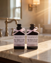 Two bottles of HGDC Essentials The Mane Event Hair Oil Treatment standing on a marble countertop in a warm, softly lit bathroom setting.