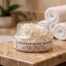 A jar of RCDC Soufflé Body Butter labeled 'Autumn After Dark' sits on a marble surface with rolled towels blurred in the background.