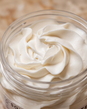 A close-up of a whipped cream or cosmetic cream swirled inside a clear glass jar, with a softly blurred warm beige background.