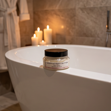 A small jar of Squebble Body Butter with Black Pepper sits on the edge of a white freestanding bathtub in a warm, candlelit bathroom setting.