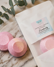 A white resealable pouch labeled 'RCDC Aromatic Shower Steamer Creativity (Grapefruit & Tangerine)' sits on a marble surface beside two round pink bath bombs, with eucalyptus leaves in the background.