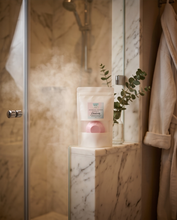 A white stand-up pouch labeled 'Bath & Body Essentials' sits on a marble ledge in a steamy bathroom setting, with a pink bath bomb visible inside and a small eucalyptus sprig beside it.