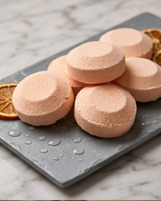 Six round, pale pink bath bombs arranged on a dark gray stone plate with water droplets, garnished with dried orange slices on a white marble surface.