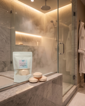 A bathroom scene with a glass shower enclosure, warm ambient lighting, marble walls, and a pouch of bath salts placed on the marble counter beside three round bath salts. A robe hangs on the right, and the shower head is visible in the background.