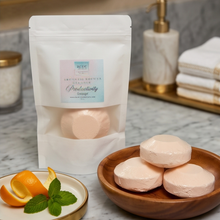 A white stand-up pouch labeled 'Productivity Grooming' stands on a marble bathroom counter beside a wooden bowl of pale pink bath bars and a small dish with orange slices and mint. A soap dispenser and folded towels are blurred in the background.
