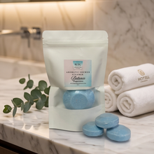A white resealable pouch labeled 'Mistral Aromatique Shower Premier Balance' stands on a marble countertop, with three round light blue bath salts in front and rolled white towels behind it in a bathroom setting.