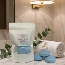 A white resealable pouch labeled 'Aromatic Shower Scrubber' stands on a marble surface beside a rolled white towel embroidered with 'HOTEL' and several round light blue bath scrubbers. Greenery and a lit wall sconce create a spa-like setting.