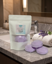 A white resealable pouch labeled 'Aromatic Shower Stones' sits on a marble bathroom counter beside three round lavender-colored shower stones, with a folded white towel and lavender sprigs nearby.
