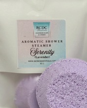 RCDC Serenity (Lavender) aromatic shower steamer in a white bag with a pastel gradient label, alongside a round lavender-colored textured steamer sponge.